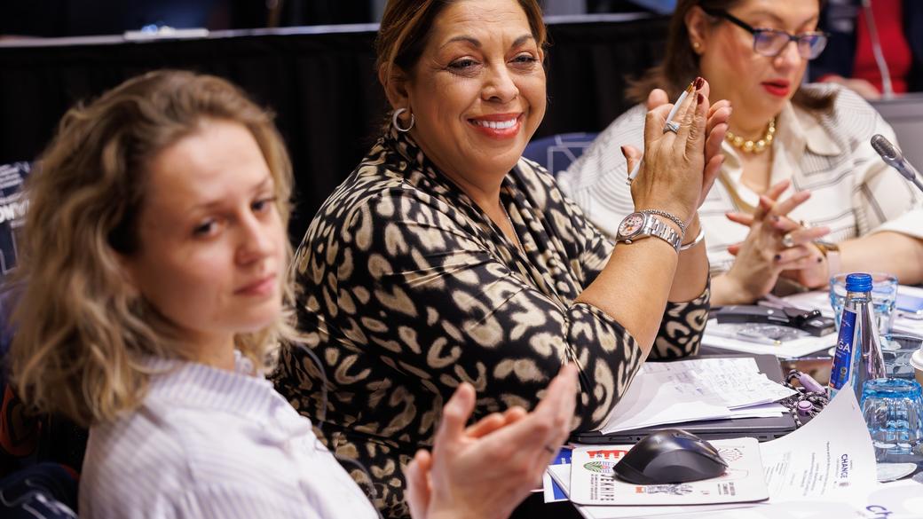 Women sitting at a conference table, smiling and clapping, with papers and water bottles in front of them.