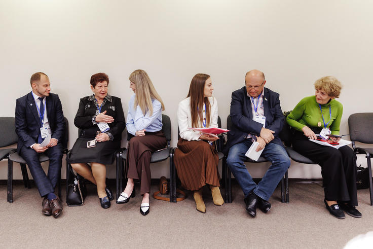 Seven people sitting in a row, engaged in conversation, wearing business attire in a meeting room.
