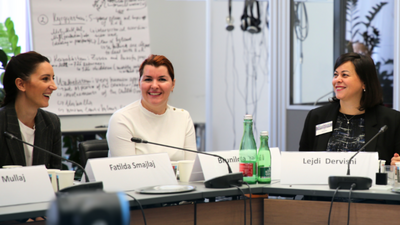 Three women smiling and talking at a conference table, with nameplates and microphones, in a bright meeting room.