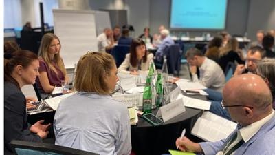 People seated around tables in a conference room, discussing and taking notes.