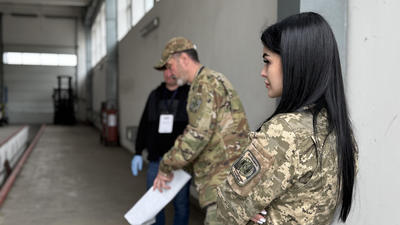 Three people in military uniforms stand in a warehouse.