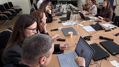 People seated at a conference table with laptops, engaging in discussion. Presentation on a screen in the background.